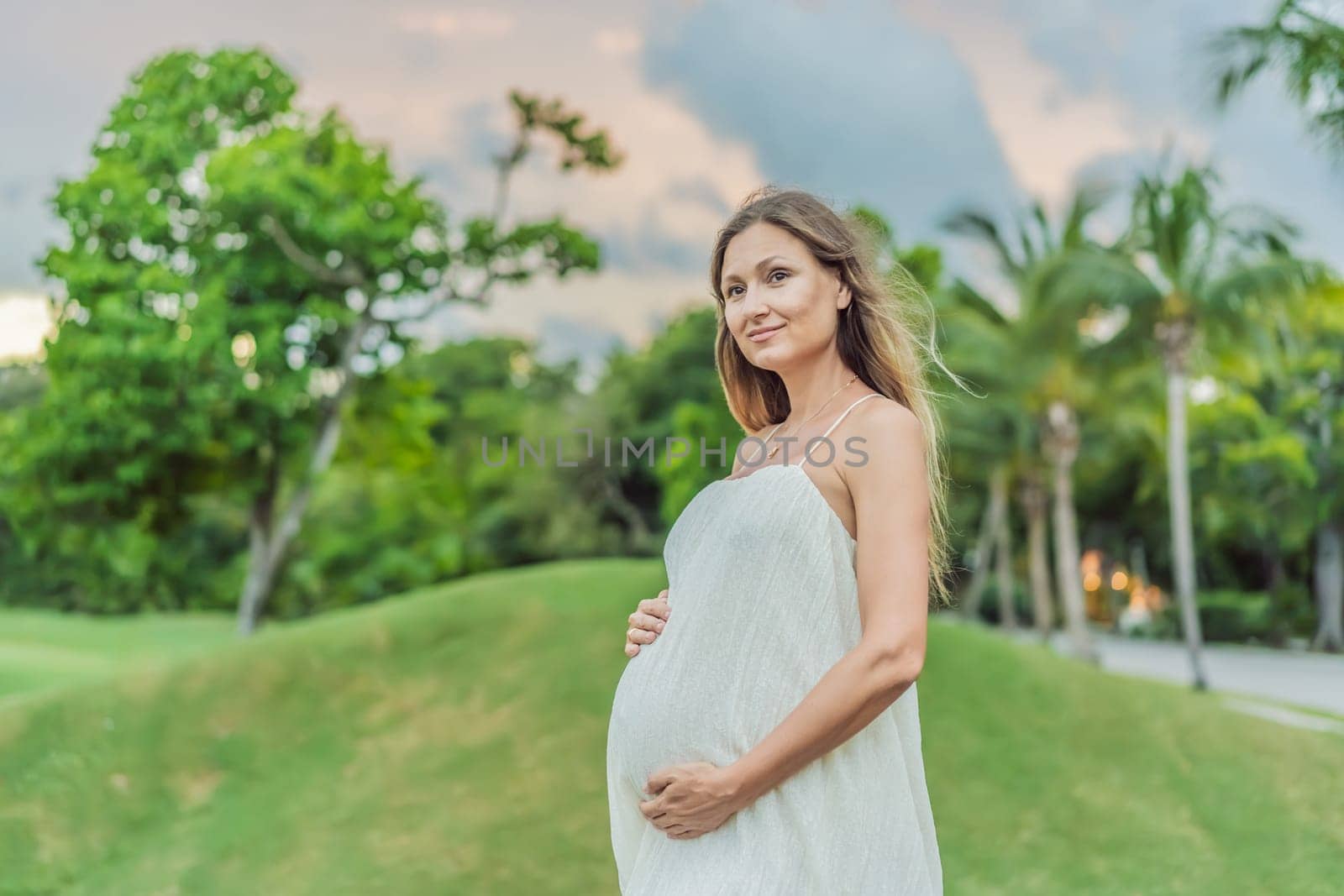 Pregnant woman hugging her tummy standing outdoors surrounded by nature. Pregnancy, expectation, motherhood concept.