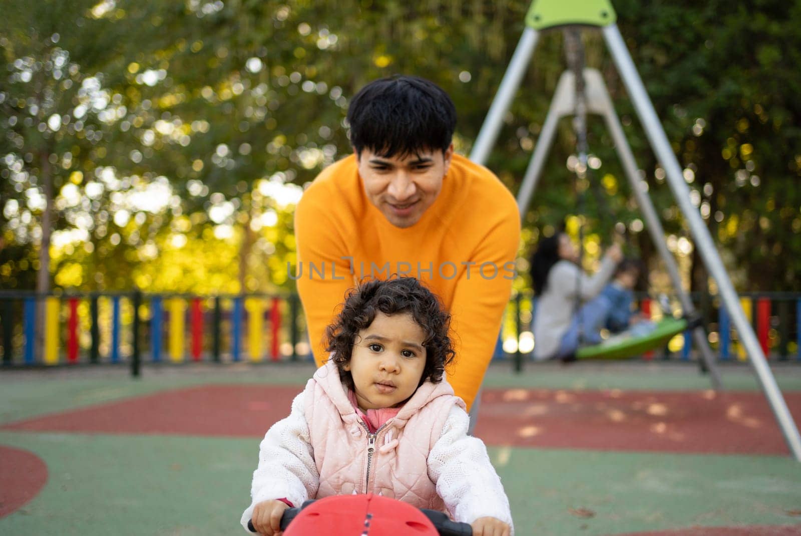 Young adult father pushing baby daughter on toy moto in playground.