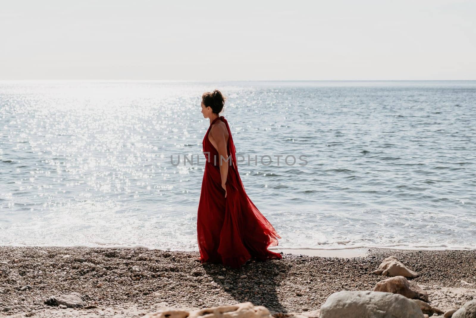 Side view a Young beautiful sensual woman in a red long dress posing on a rock high above the sea during sunrise. Girl on the nature on blue sky background. Fashion photo.