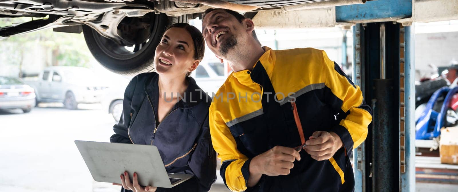 Two vehicle mechanic working together underneath lifted car, conduct car inspection with laptop. Automotive service technician in uniform carefully make diagnostic troubleshooting. Panorama Oxus