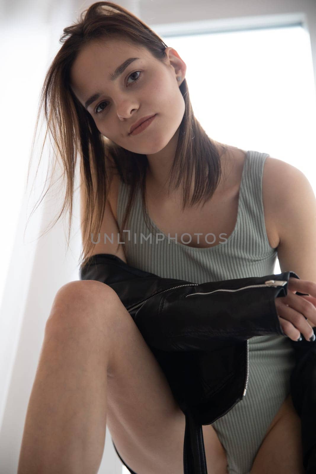 a woman in a gray bodysuit and a black jacket stands by the window