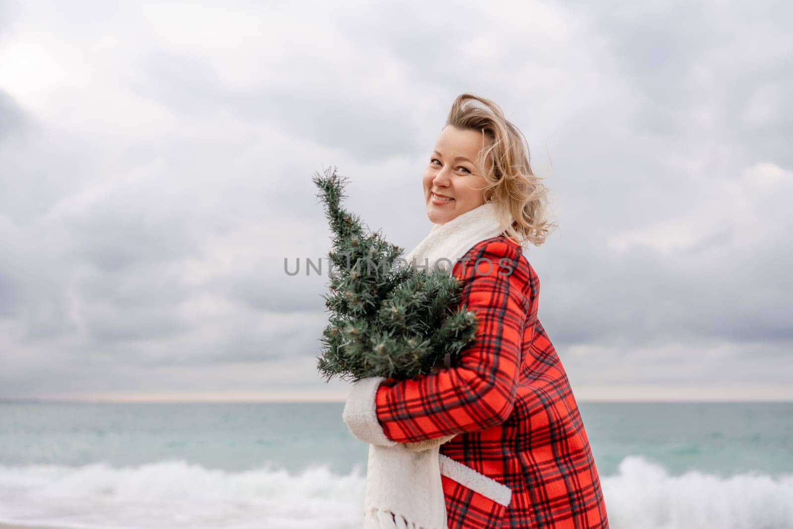 Blond woman holding Christmas tree by the sea. Christmas portrait of a happy woman walking along the beach and holding a Christmas tree in her hands. Dressed in a red coat, white dress. by Matiunina