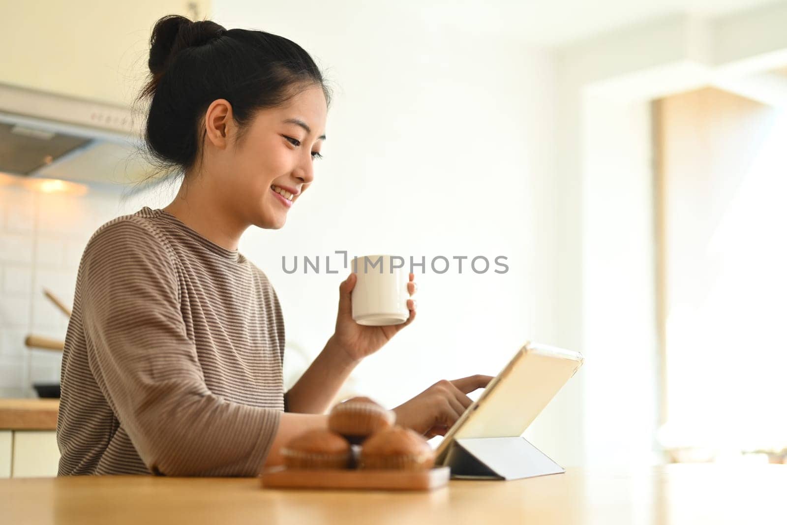 Attractive young woman drinking coffee and using digital tablet at table in kitchen.