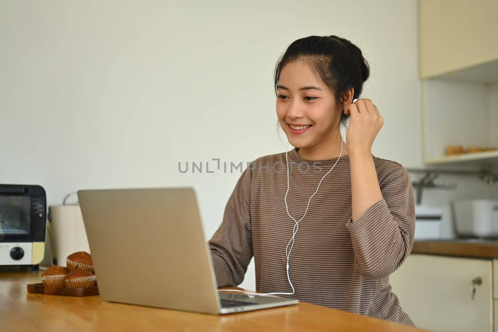 Charming young woman sitting at kitchen counter and working online or browsing internet on laptop.