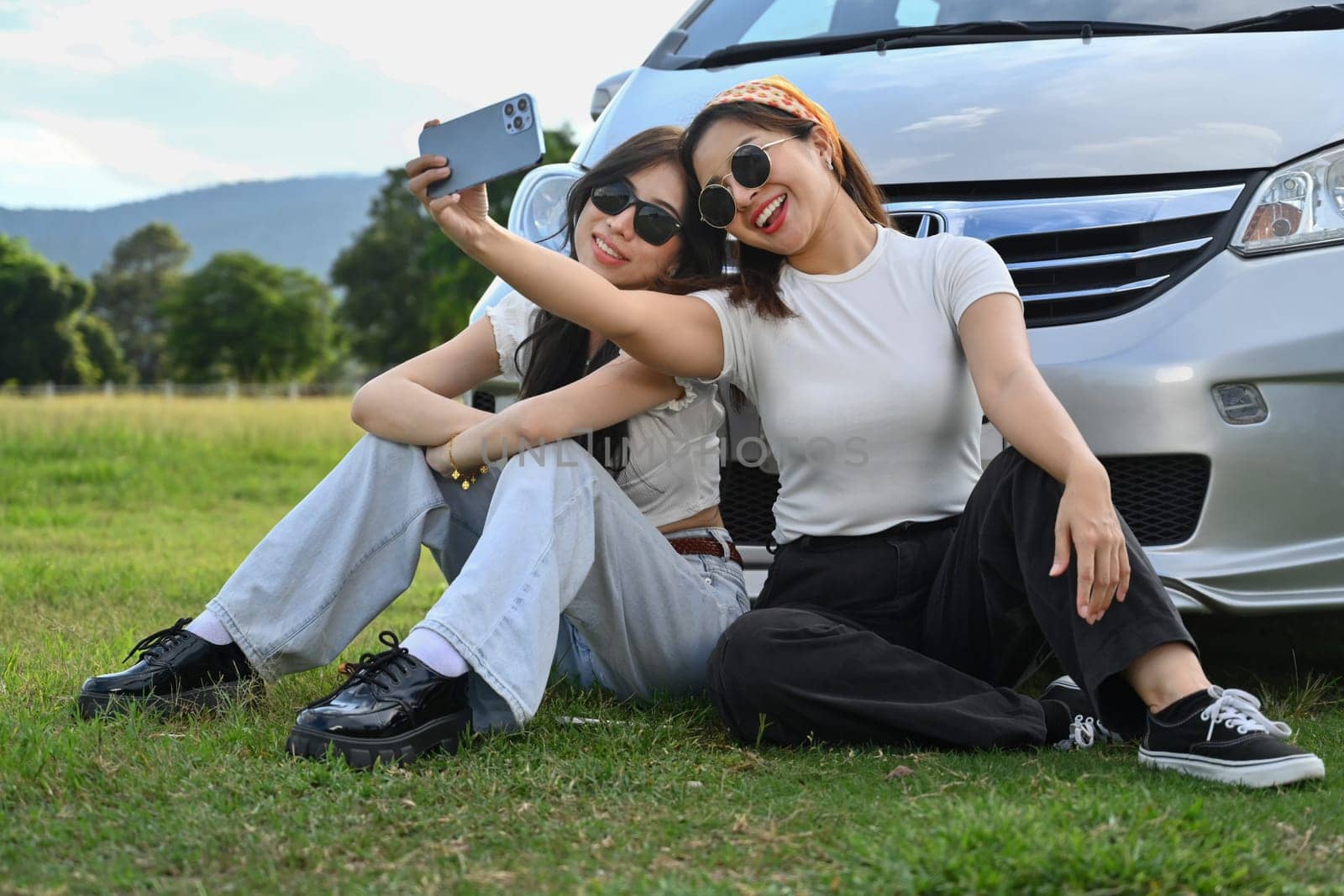 Cheerful female friends taking a break from driving siting in front of car on meadow and taking selfies with smartphone by prathanchorruangsak