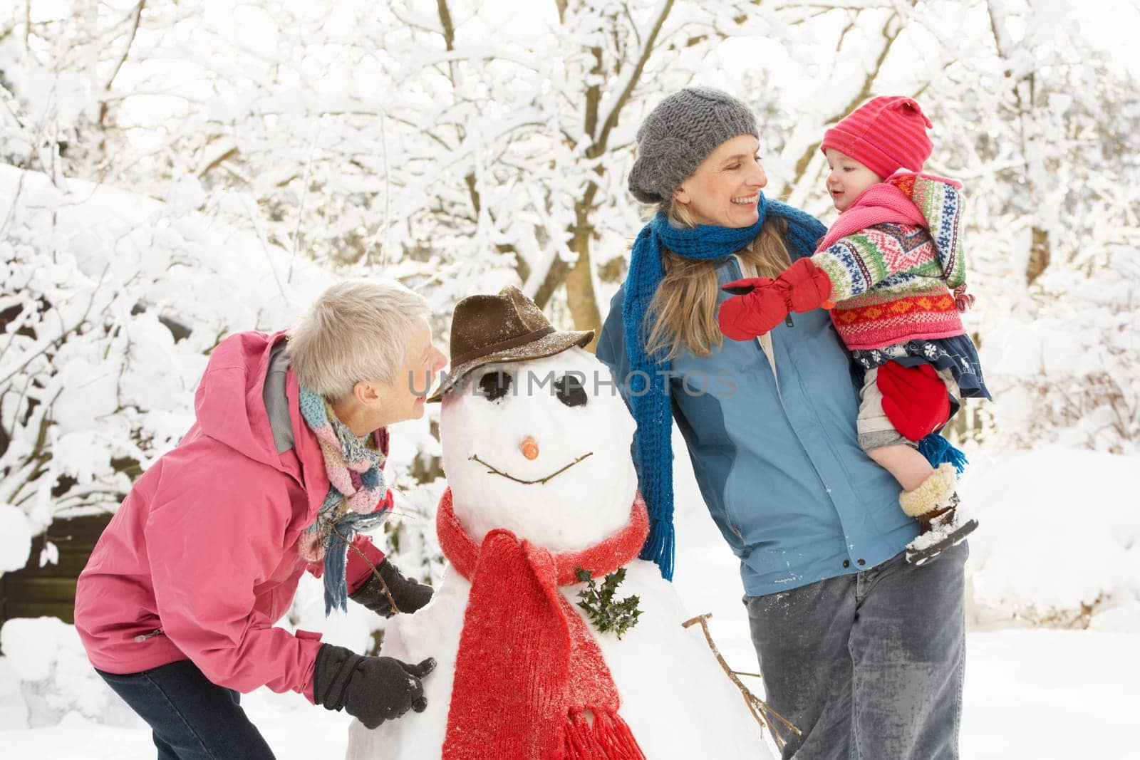 Young Girl With Grandmother And Mother Building Snowman In Garden