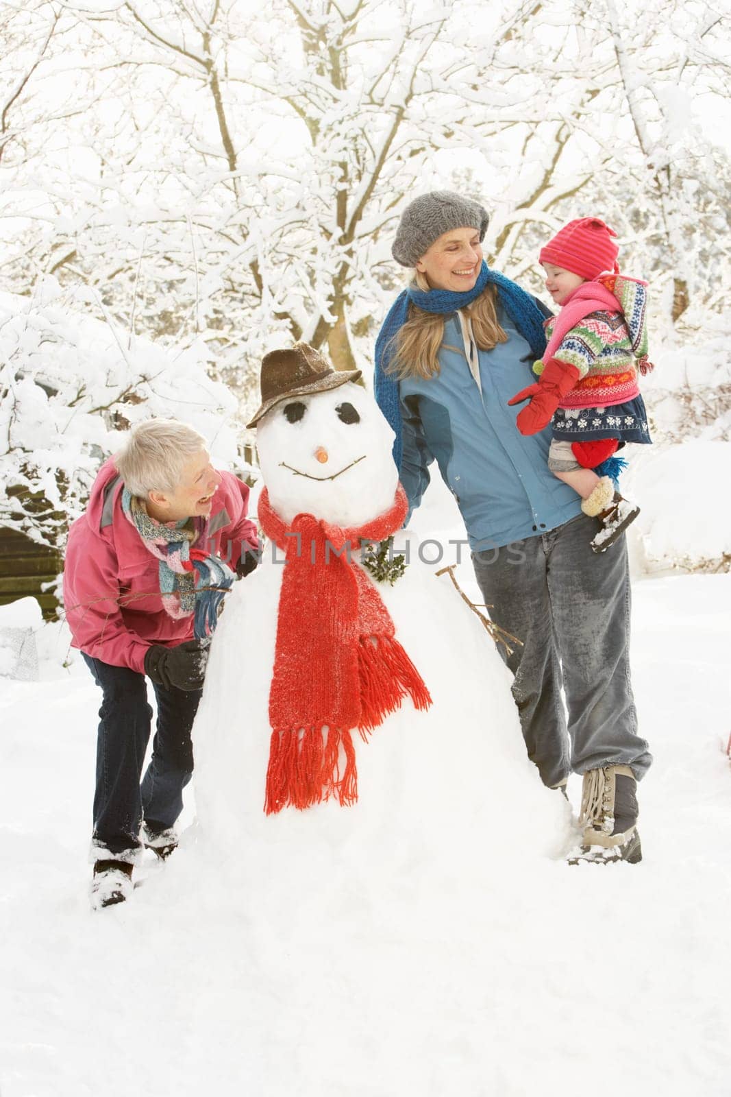 Young Girl With Grandmother And Mother Building Snowman In Garden