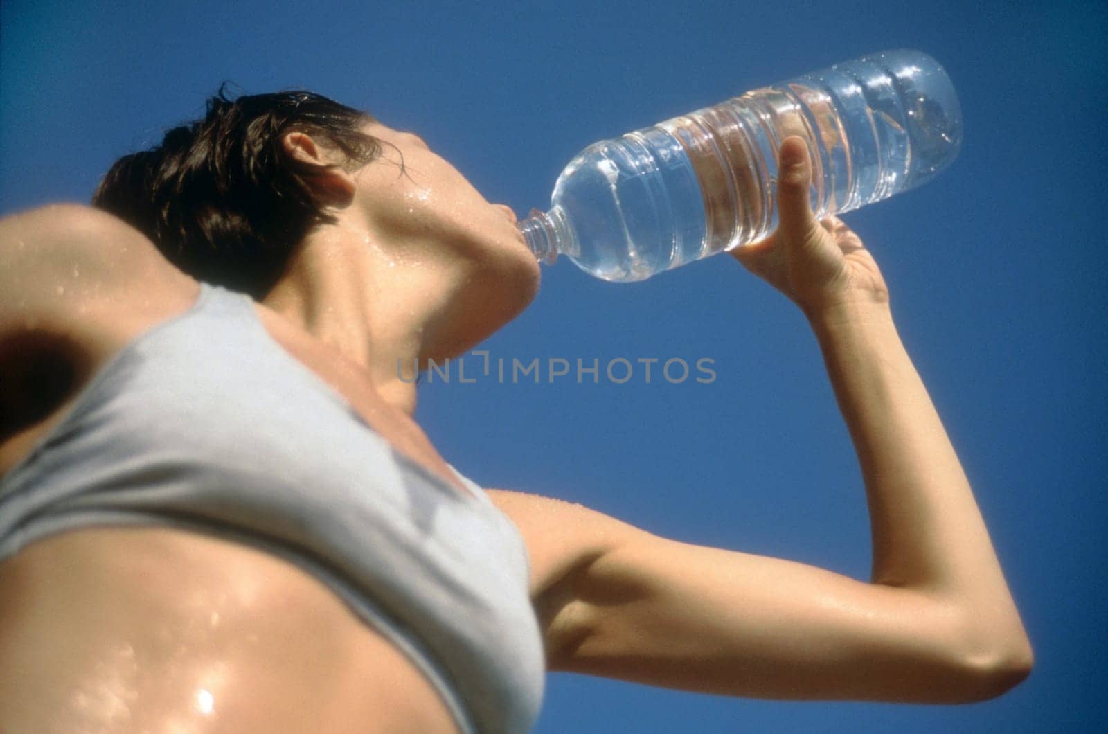 Low angle view of a woman in exercise gear, drinking water from a water bottle against a blue sky. Horizontal shot.