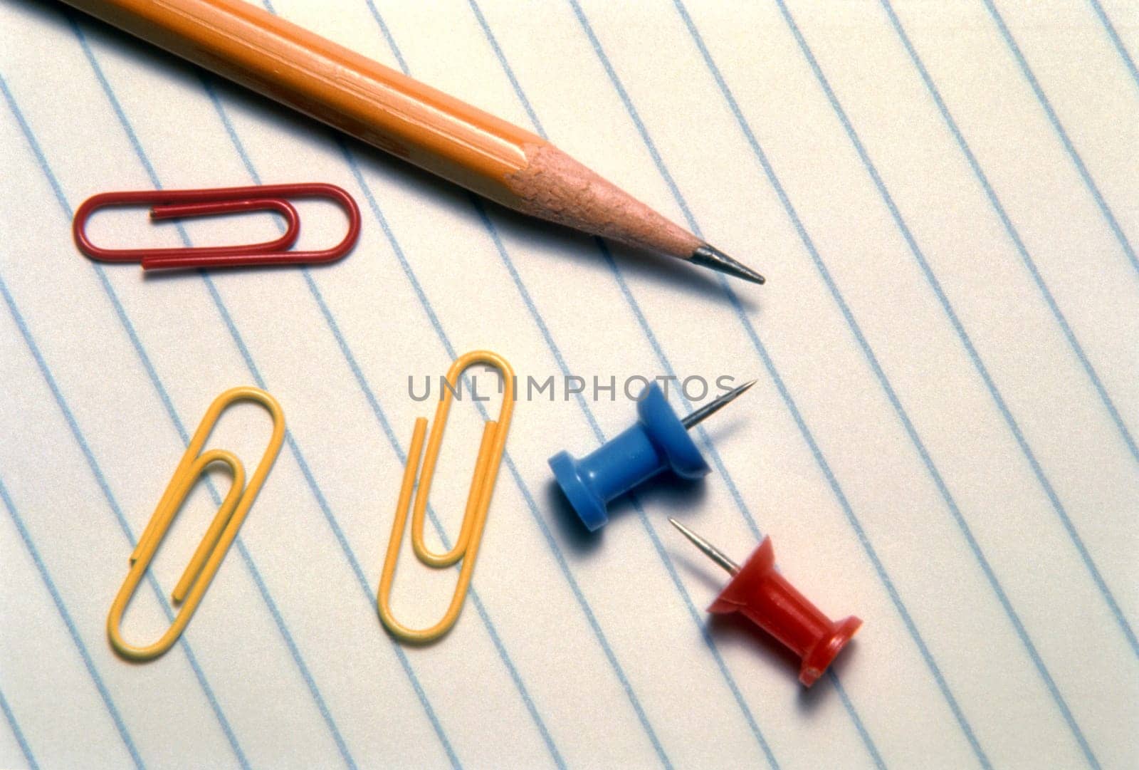 A pencil, paper clips and pushpins are lying on a piece of lined notebook paper. Horizontal shot.