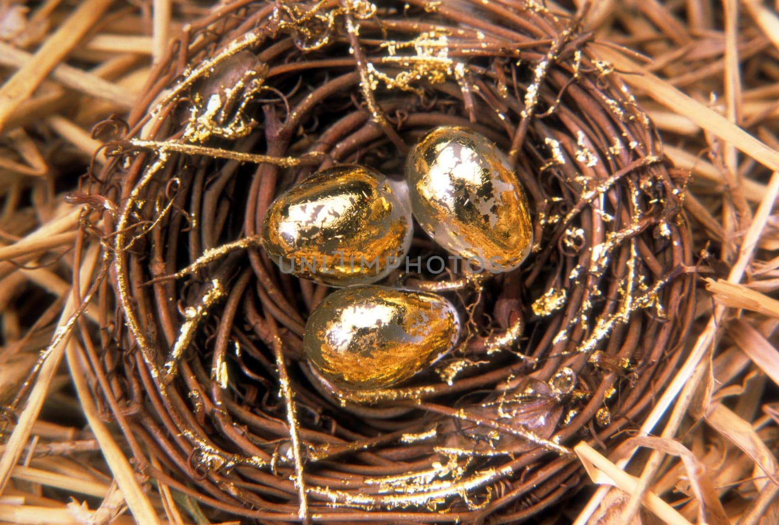 Overhead view of a woven nest sitting in straw with three golden eggs. Horizontal shot.