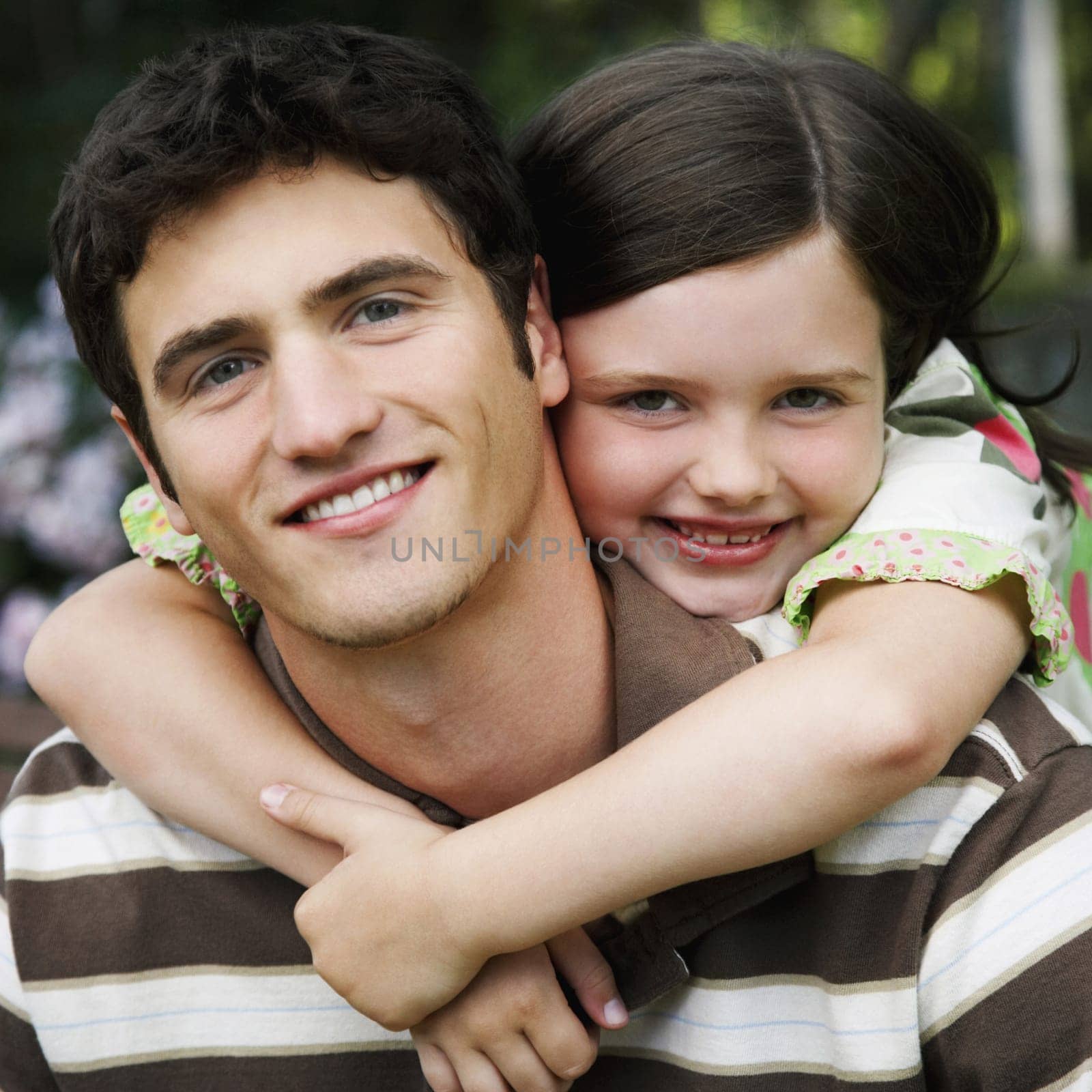 Young girl wraps her arms around a young man from behind. They are both smiling towards the camera. Square shot.