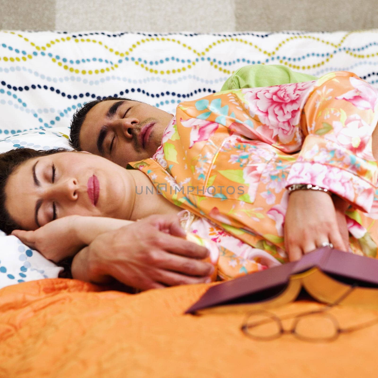 A man and woman sleep on a bed with a book and eye glasses in the foreground. Square format.