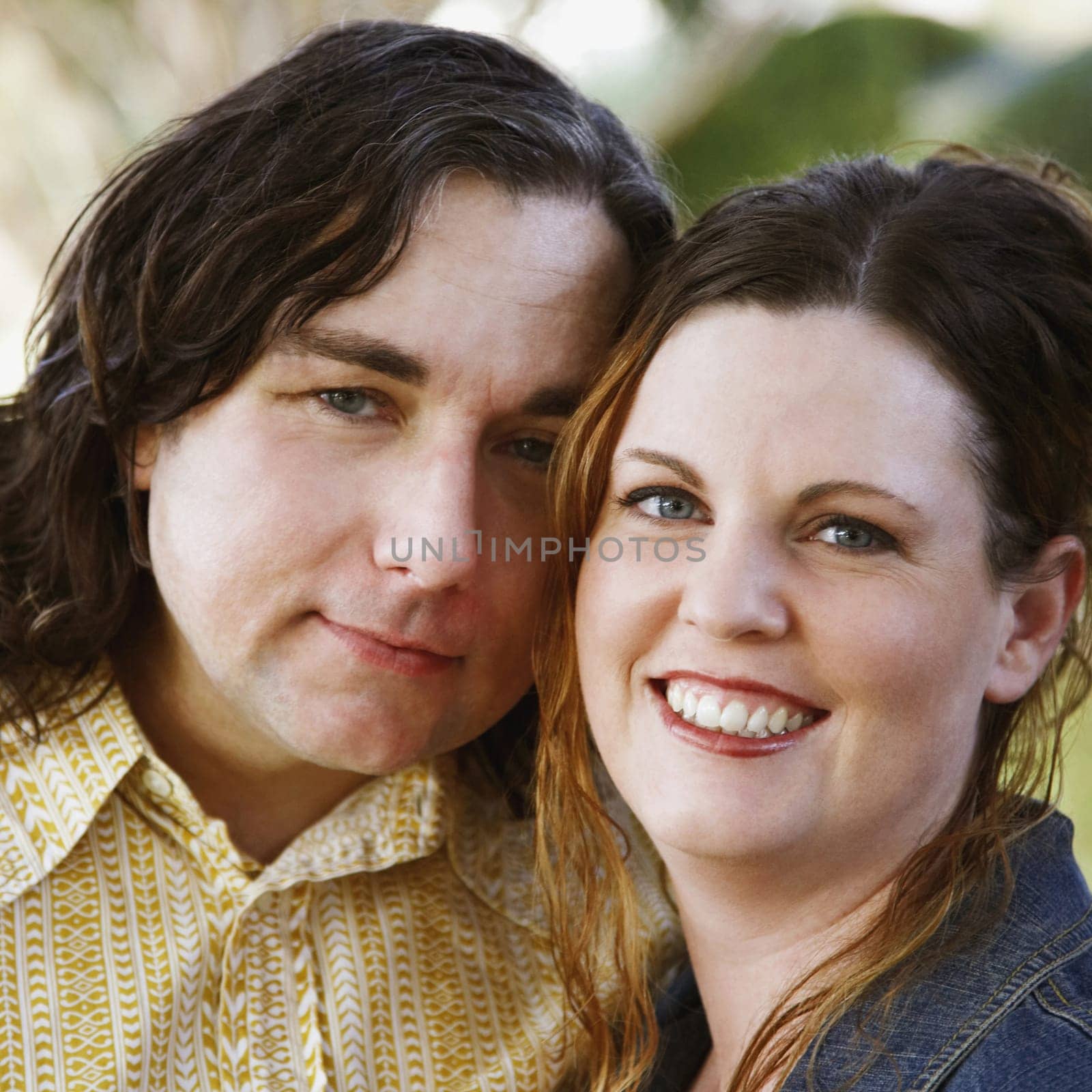 An affectionate couple with their heads leaning into each other smile towards the camera. Square shot.