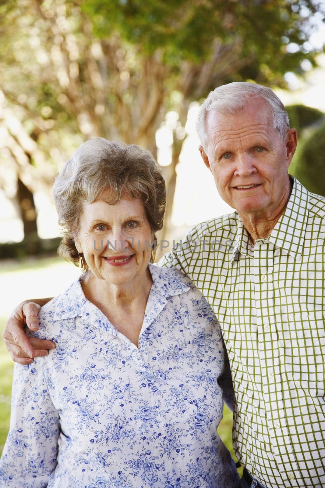 Elderly man standing with his arm around an elderly woman in their yard smiling at the camera. Vertical shot.