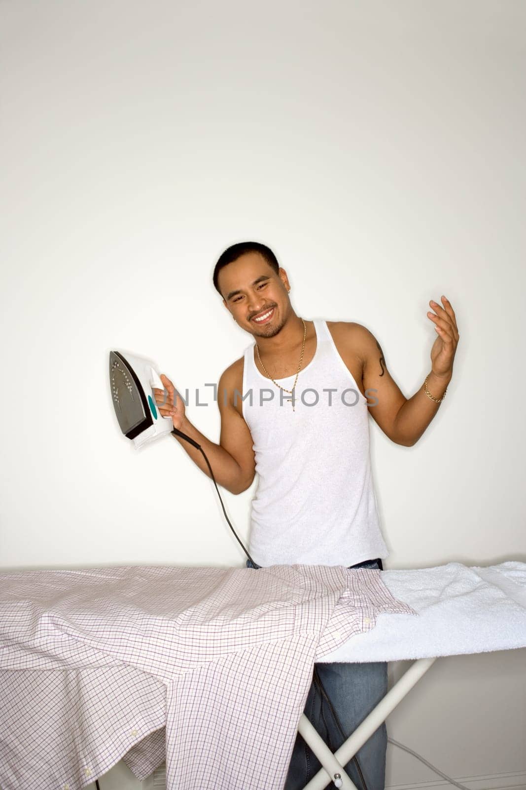 Happy young man standing behind an ironing board. He is shrugging and holding an iron. Vertical shot.