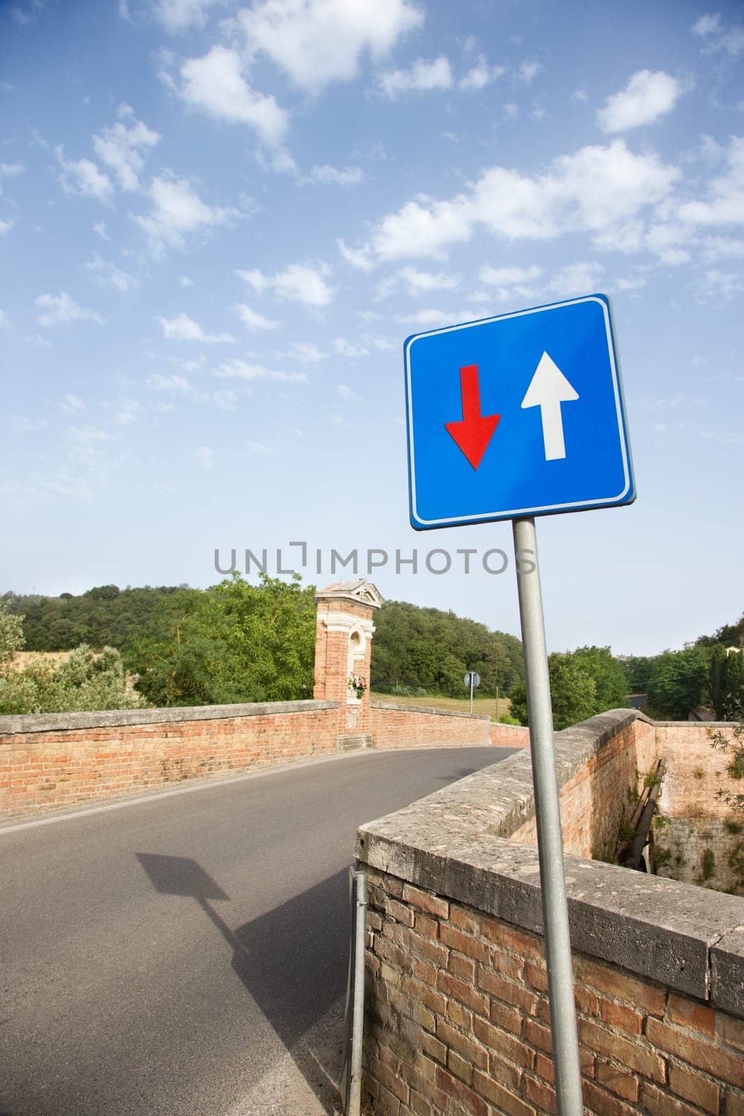 View of a rural road lined with a low brick wall.  In the foreground is a road sign with arrows. Vertical shot.