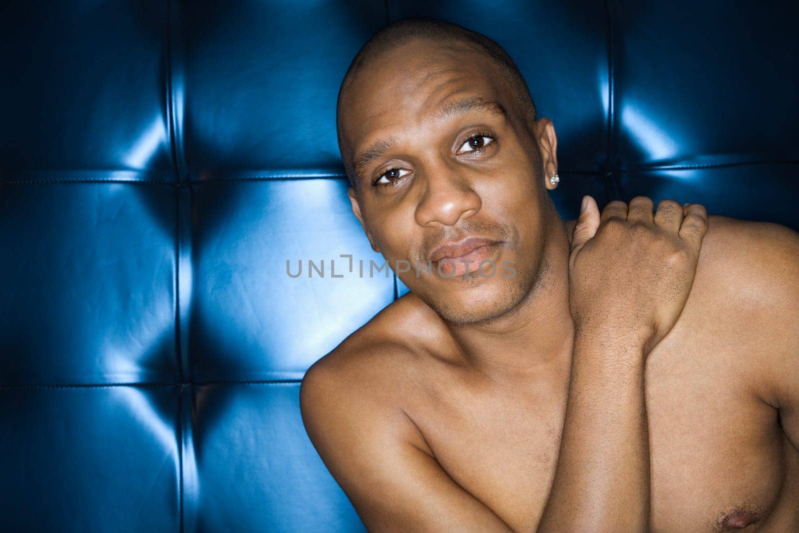 Handsome young man sitting with his hand on his shoulder against blue padded background, shirtless and smiling. Horizontal shot.