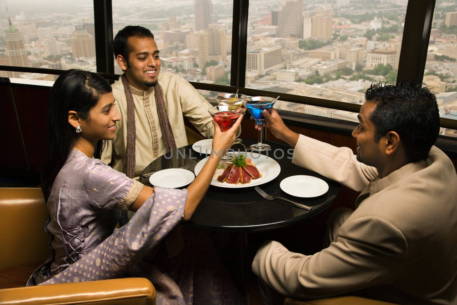 High angle view of young adult males and a female seated near a window of a high rise restaurant. They are toasting with martinis. Horizontal shot.