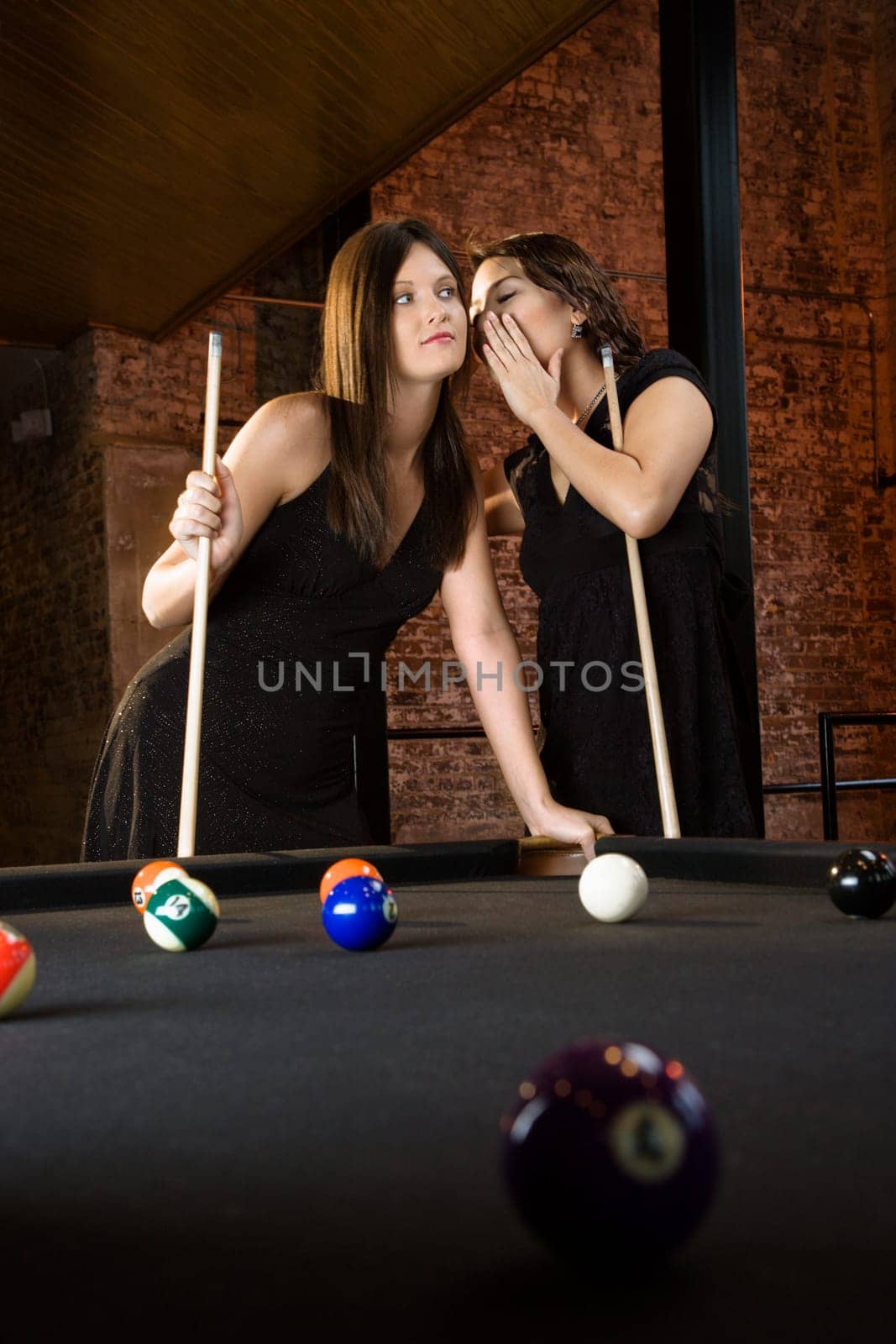 Two attractive young women at pool table as one whispers into other's ear.