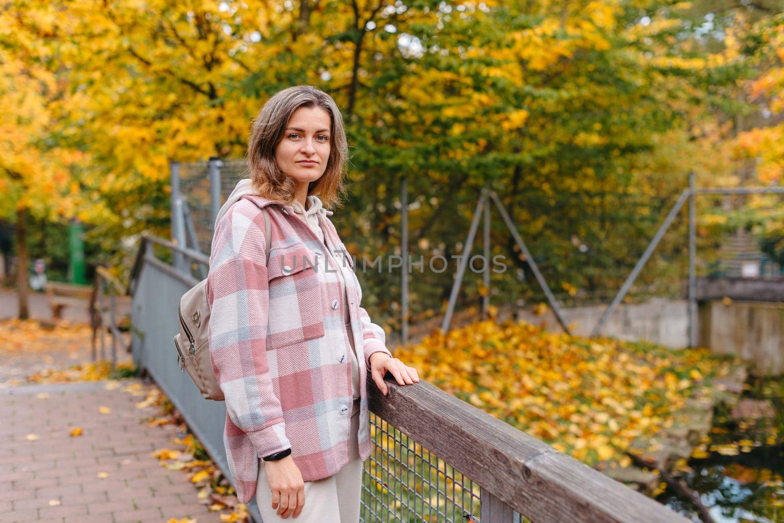 Portrait of cute young woman in casual wear in autumn, standing on bridge against background of an autumn Park and river. Pretty female walking in Park in golden fall. Copy space. smiling girl in the park standing on wooden bridge and looking at the camera in autumn season