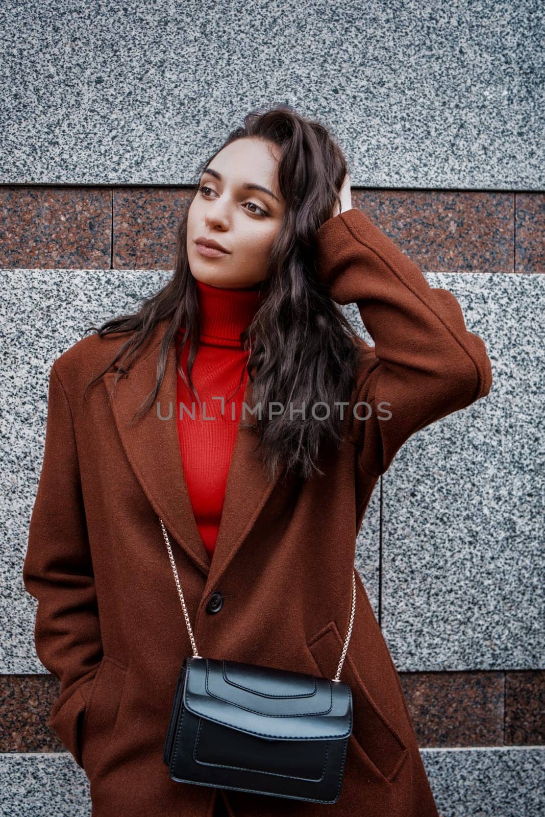 fashionable young woman in a brown coat and red sweater poses against the wall