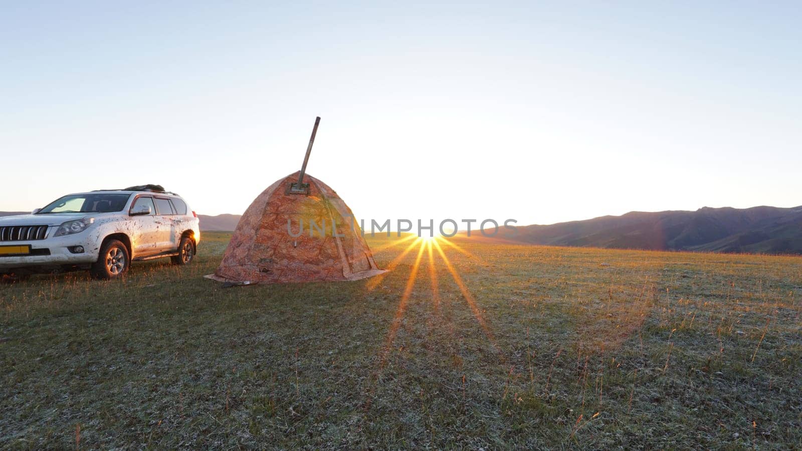 Sunrise among green fields and hills. Camping. Camping. There is a tent, there is smoke from the stove. There is a white SUV car nearby. The sun rays come out from behind the horizon. Kazakhstan