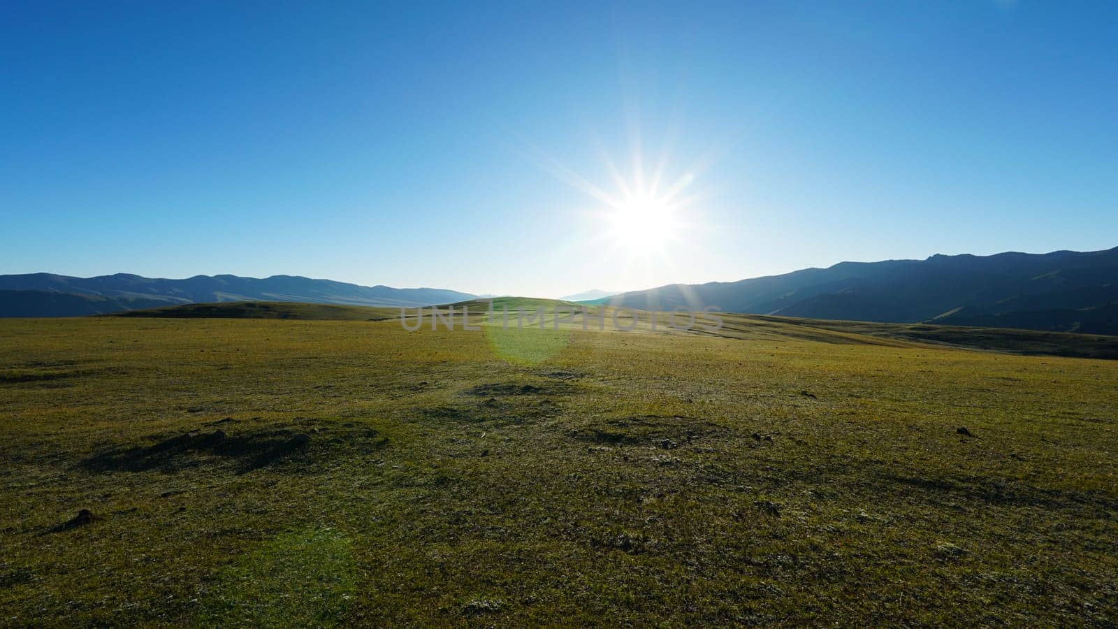 Sunrise with a view of green hills and snowy peaks. Spacious green fields and yellow grass in places. Blue clear sky. Shadows from mountains, gorges. Peace and relaxation. Outdoor recreation
