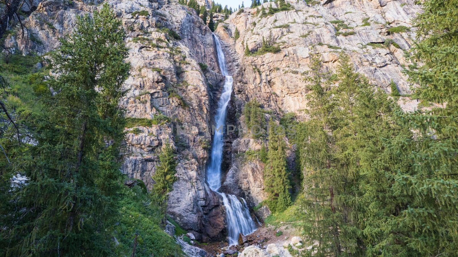 A high waterfall cascades in a mountain gorge. Coniferous trees, Christmas trees and grass grow. The river runs. Hiking trails. Top view from a drone. Sunset. Burkhan Bulak Waterfall, Kazakhstan