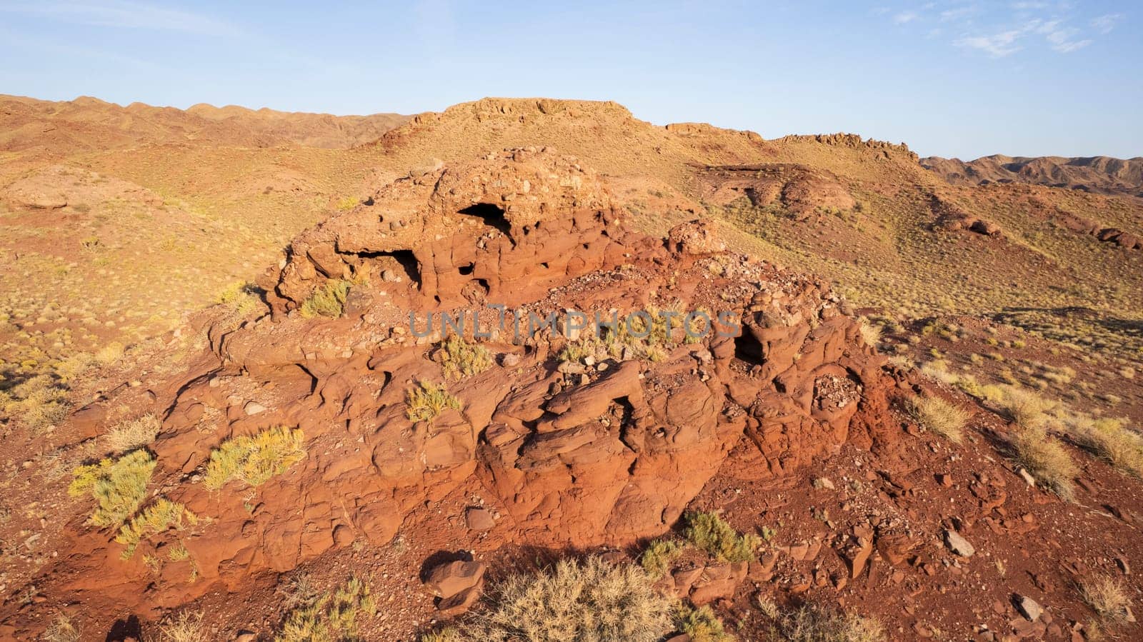 A volcanic place. Frozen old lava. Red Mountains. The rock is red-maroon in color, caves were formed in the rocks. Grass and bushes grow. The view from the drone. An unborn volcano. Kazakhstan.