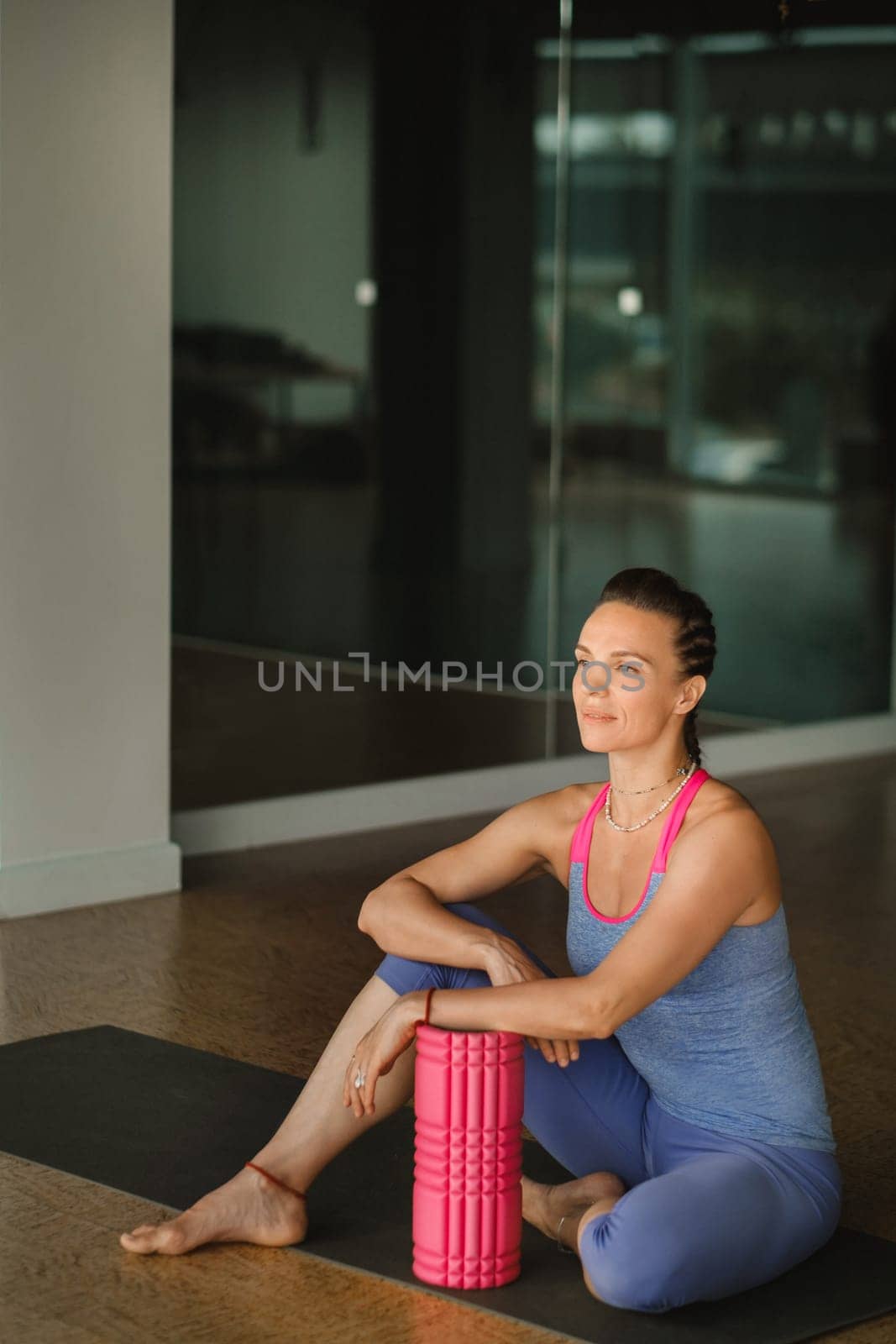 A slender girl is sitting on a yoga mat with a big massage roller in the fitness room.