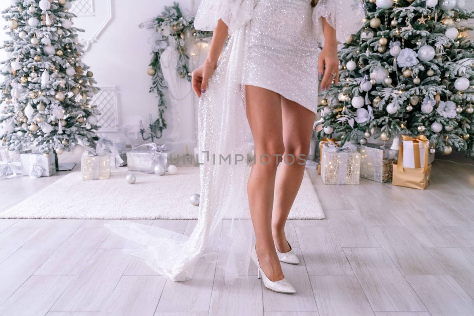 Women legs christmas tree. Cropped photo of beautiful elegant female legs in white high-heeled shoes and a white short dress, a woman stands against the background of a Christmas tree with gifts by Matiunina