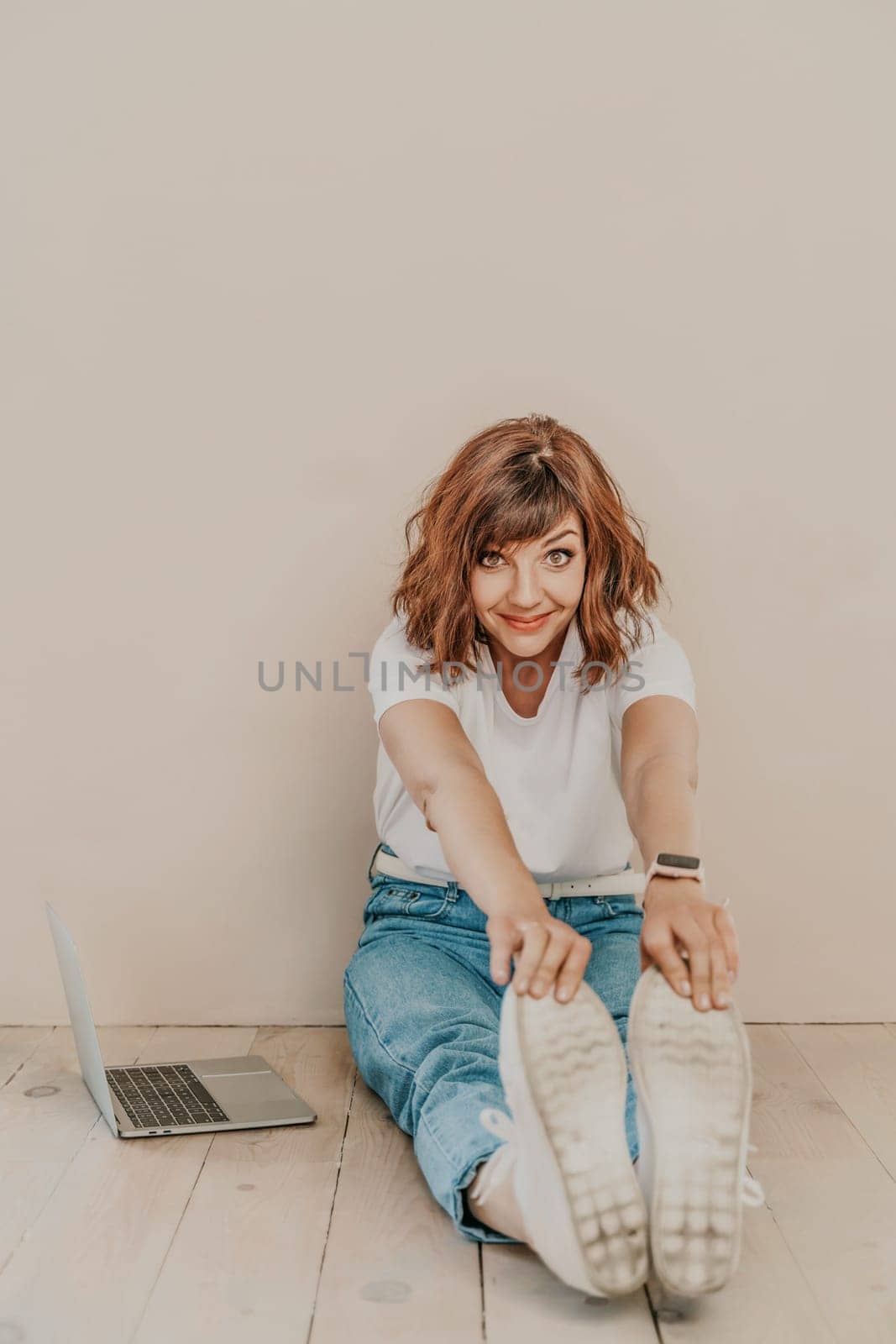 A brunette sits on the floor with a laptop on a beige wall background. She is wearing a white T-shirt, jeans and white sneakers