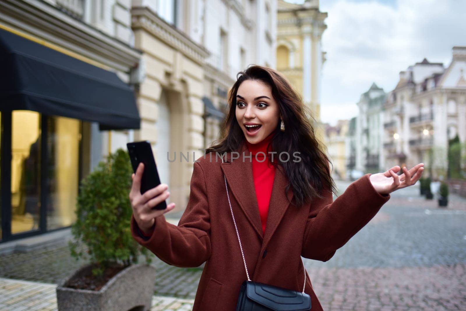 A young woman in a red coat walks down the street and experiences joyful emotions looking at the smartphone in her hand. Good News, Winning, Discounts