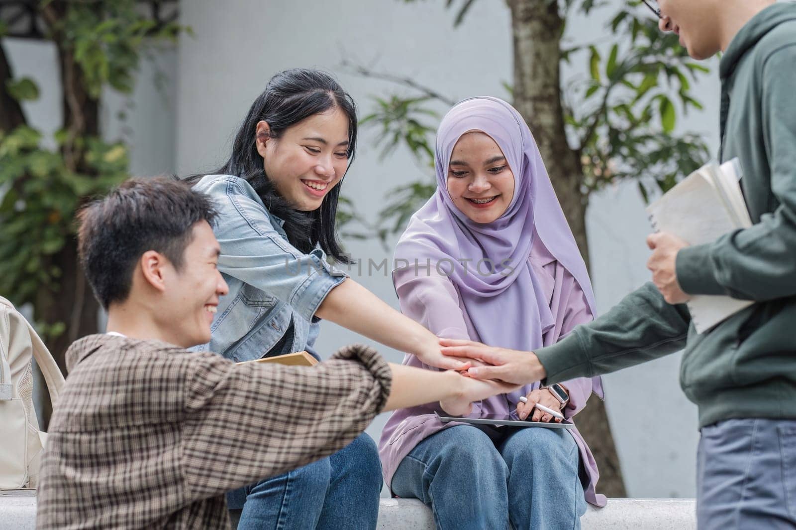 Image of a group of cheerful Asian college students putting their hands together, showing teamwork and unity. by wichayada