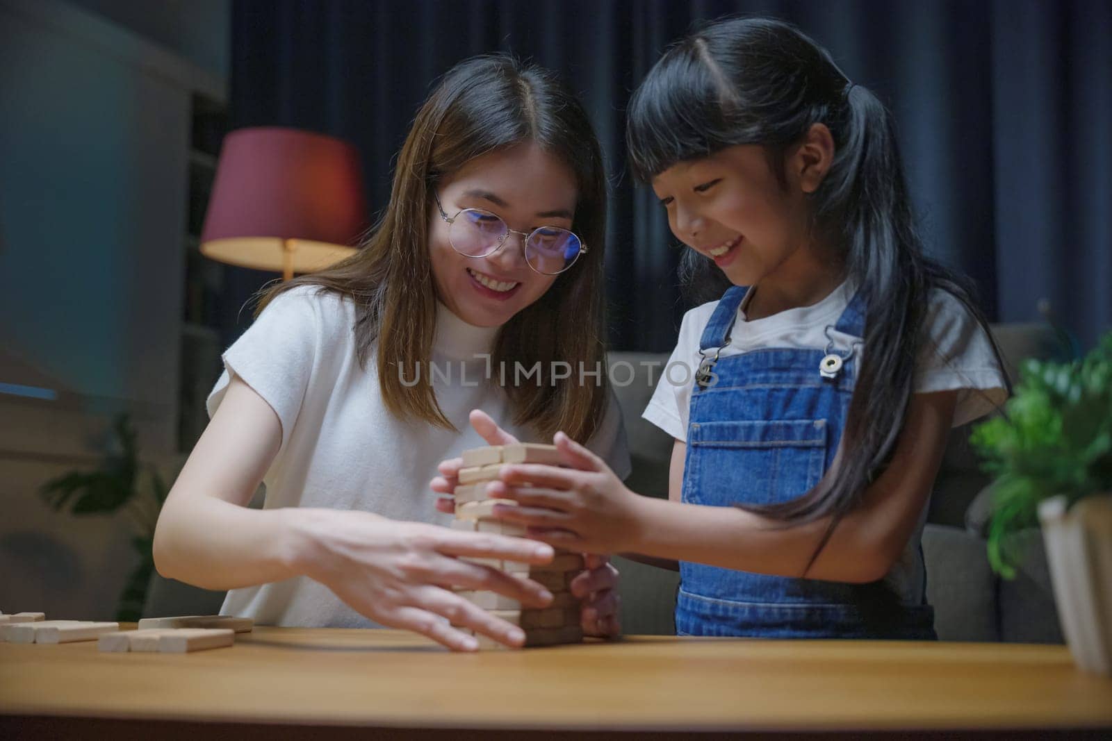 Asian young mother playing game in wood block with little daughter in home living room at night, Smiling woman help teach preschooler kid play build constructor tower of wooden blocks, family funny
