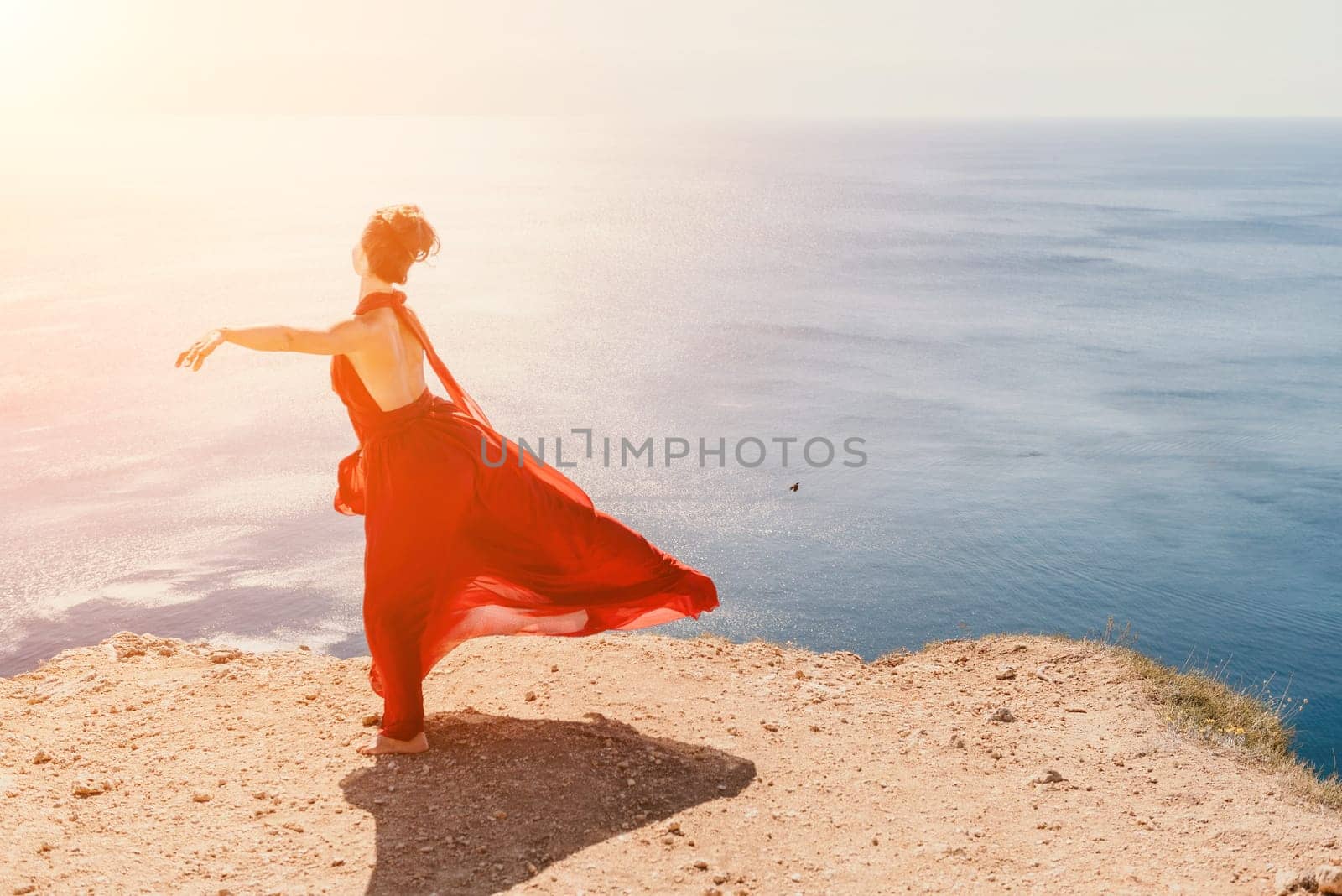 Side view a Young beautiful sensual woman in a red long dress posing on a rock high above the sea during sunrise. Girl on the nature on blue sky background. Fashion photo.