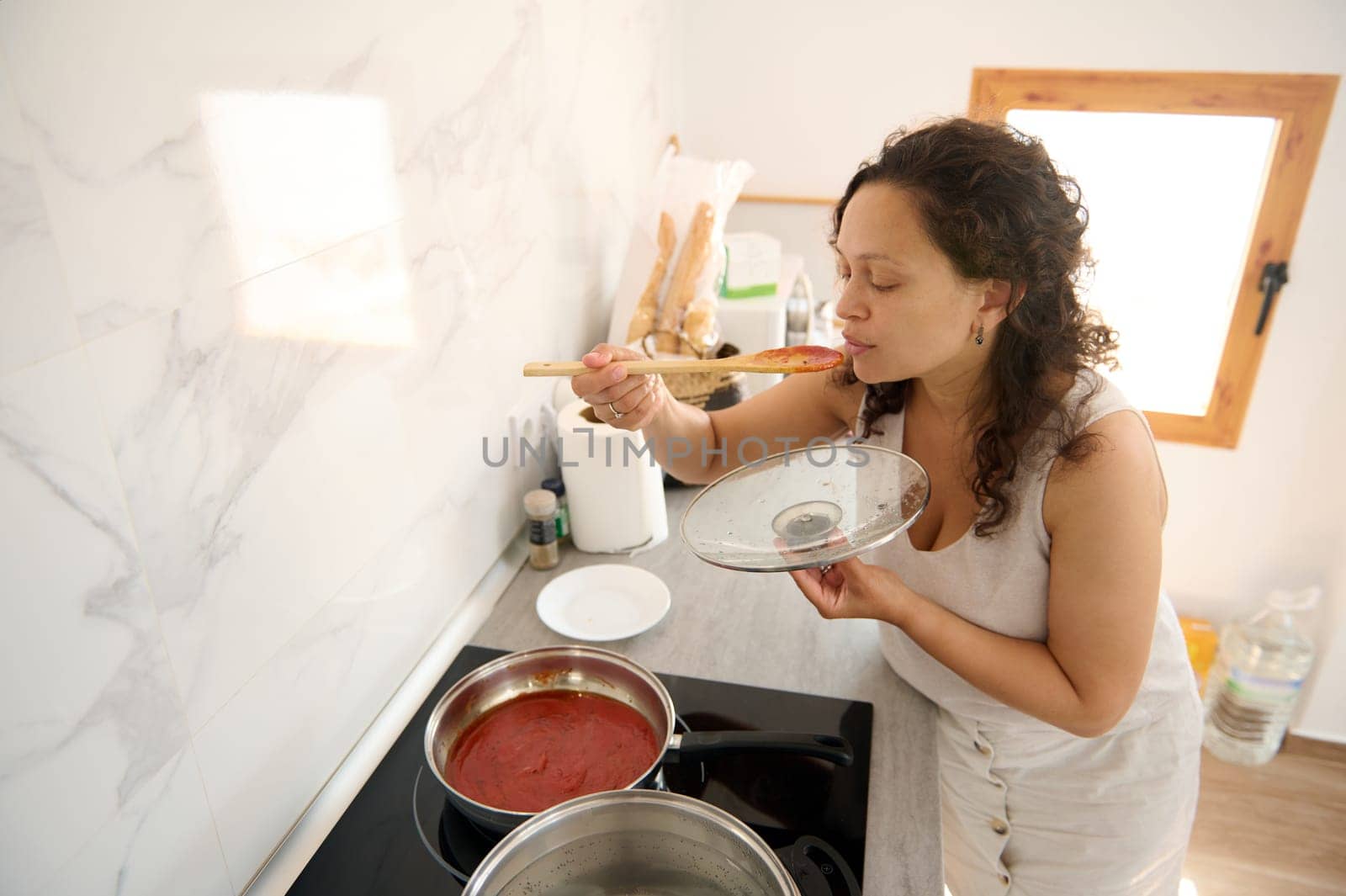 Young Latin American pretty housewife tasting the tomato sauce she's cooking in the home kitchen. People. Culinary. Lifestyle