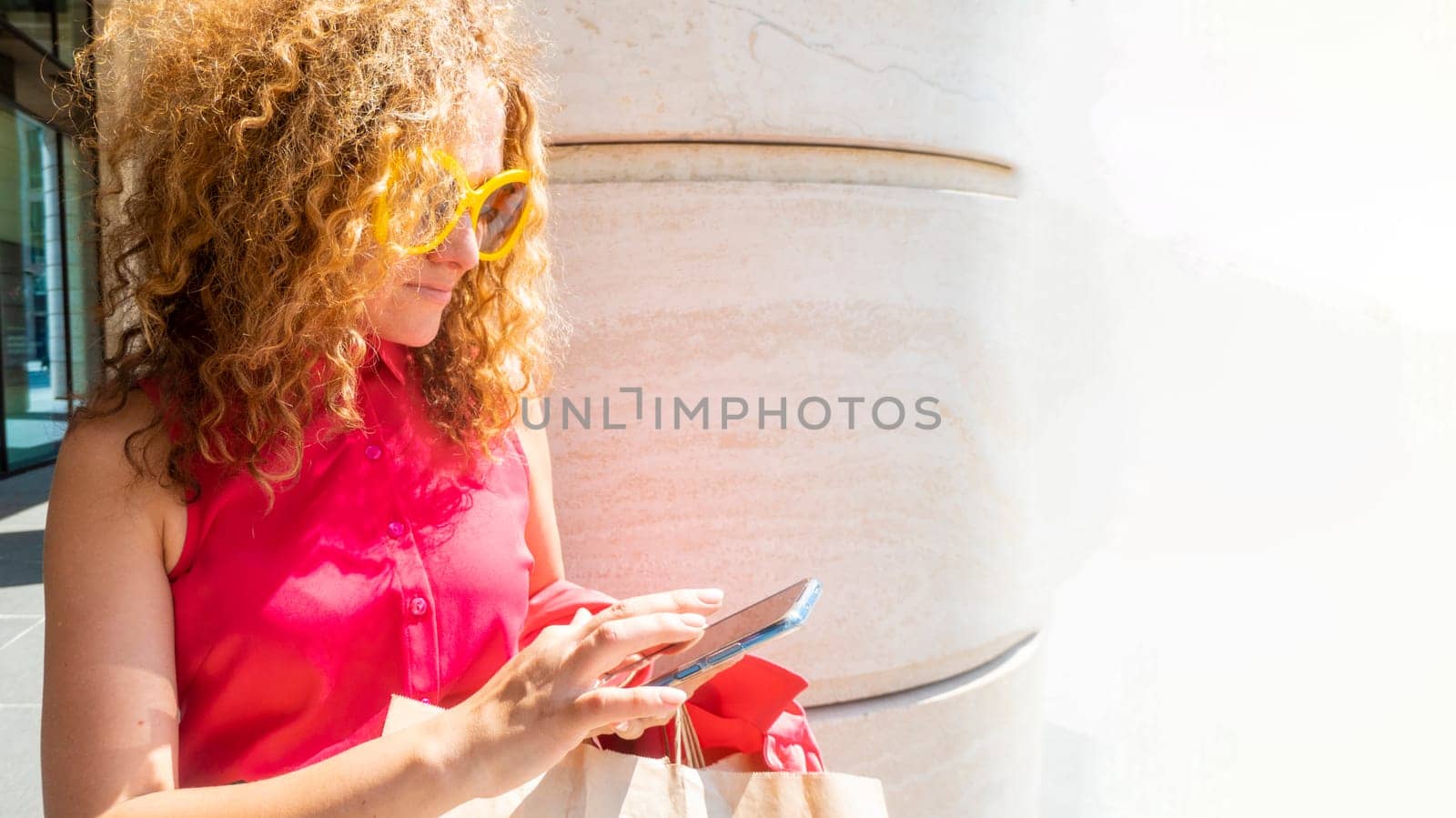 Coquettish s young female model in fashionable elegant bright multicolored green and red clothes looks at her phone against the background of the columns of an office building in the city. the concept of mobile communication, remote work, online shopping