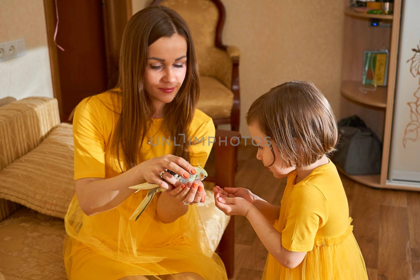 Sweet mommy and cute daughter girl with a parrot in yellow dresses in the room by jovani68