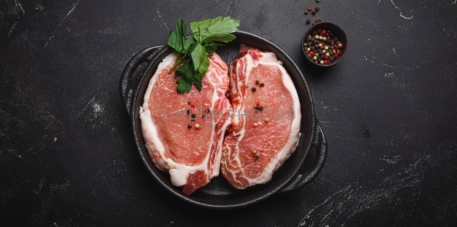 Cut raw meat pork steaks with seasonings in black cast iron pan on dark rustic stone background top view, ready for roasting. Pork loin chops cooking by its_al_dente