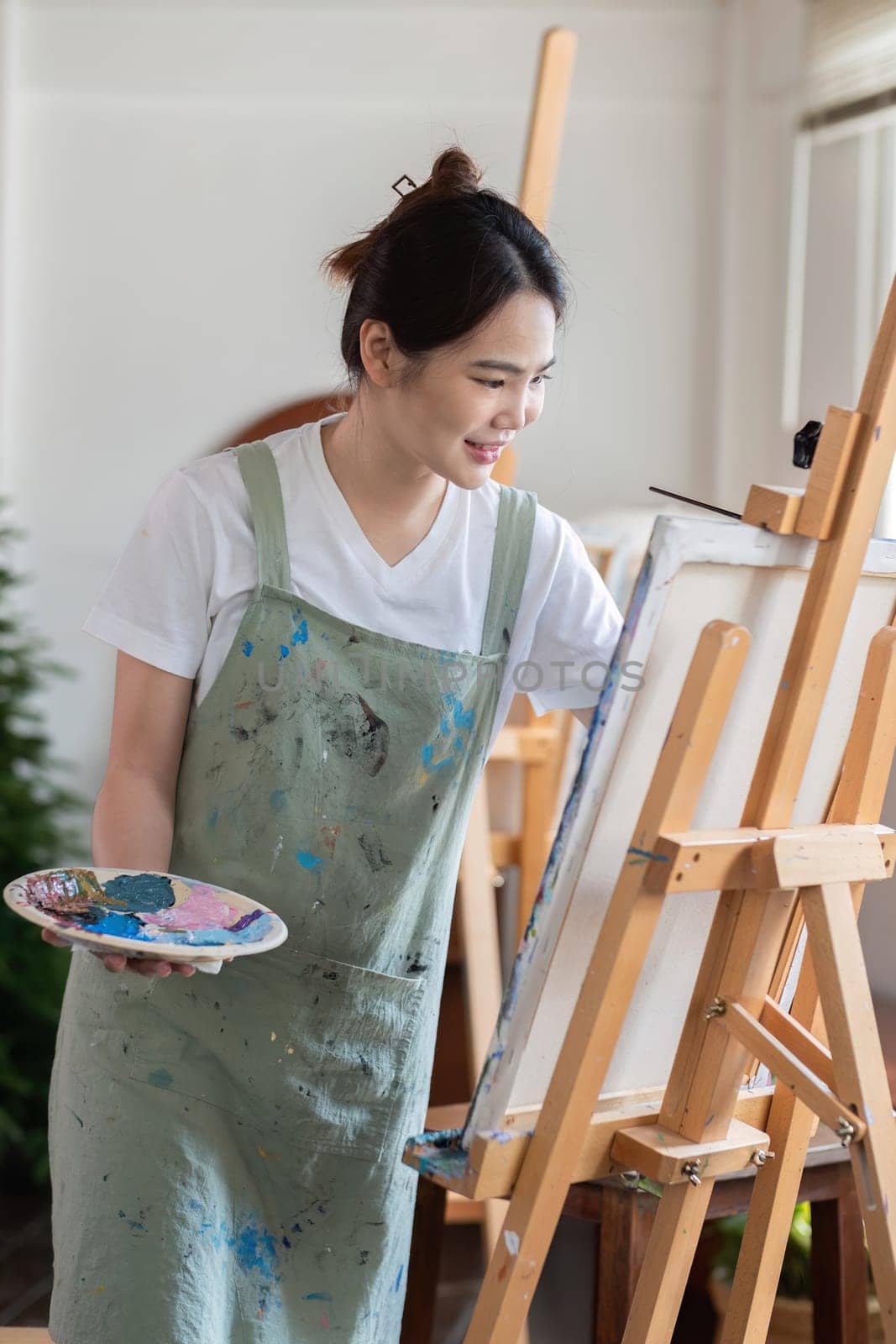 Attractive young Asian woman concentrates on painting with acrylic paints on canvas in painting studio. with a cheerful smile.