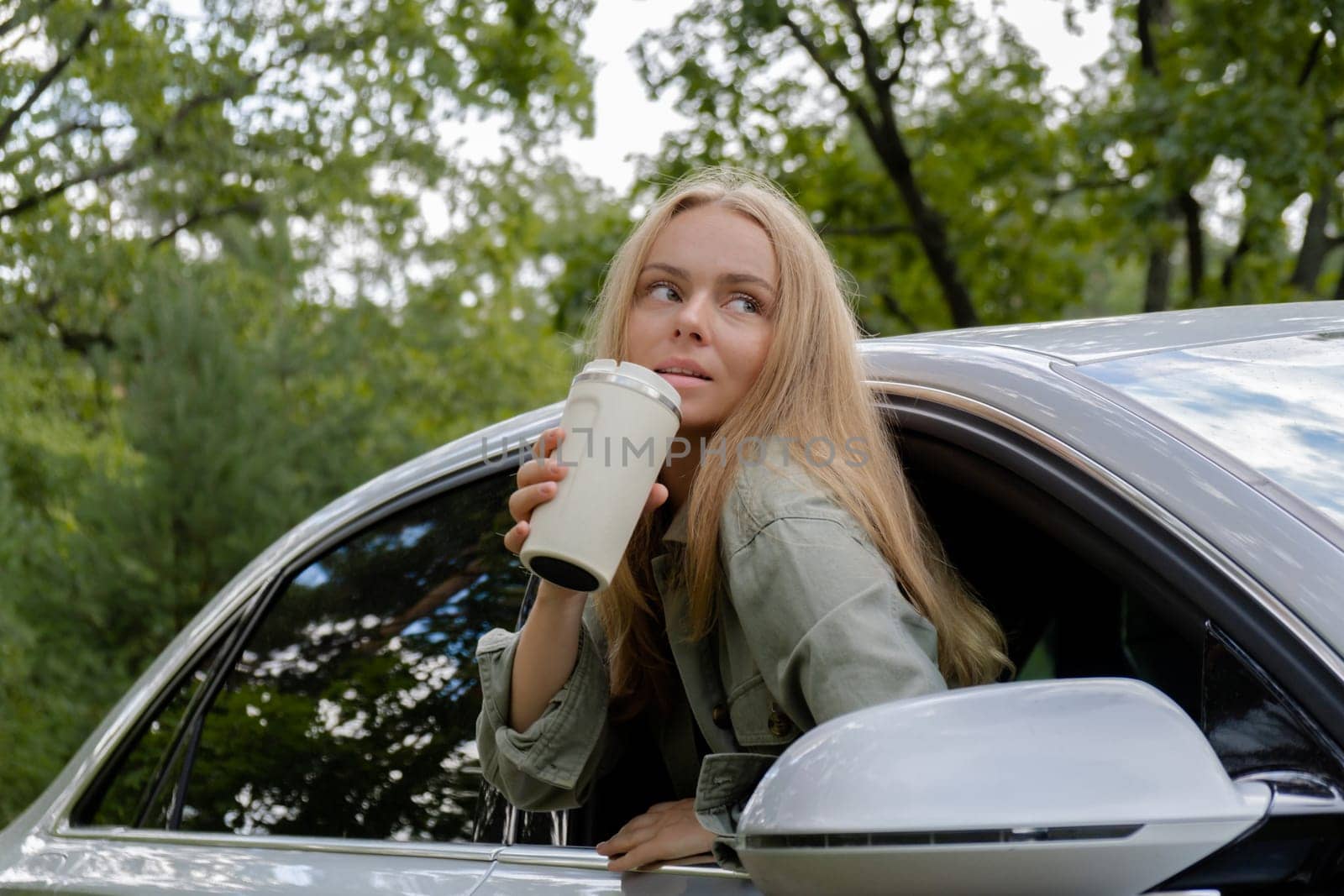 Smiling young woman looking from car window and drinking coffee or tea from reusable thermos cup. Local solo travel on weekends concept. Exited woman explore freedom outdoors in forest. Unity with nature lifestyle, rest recharge relaxation