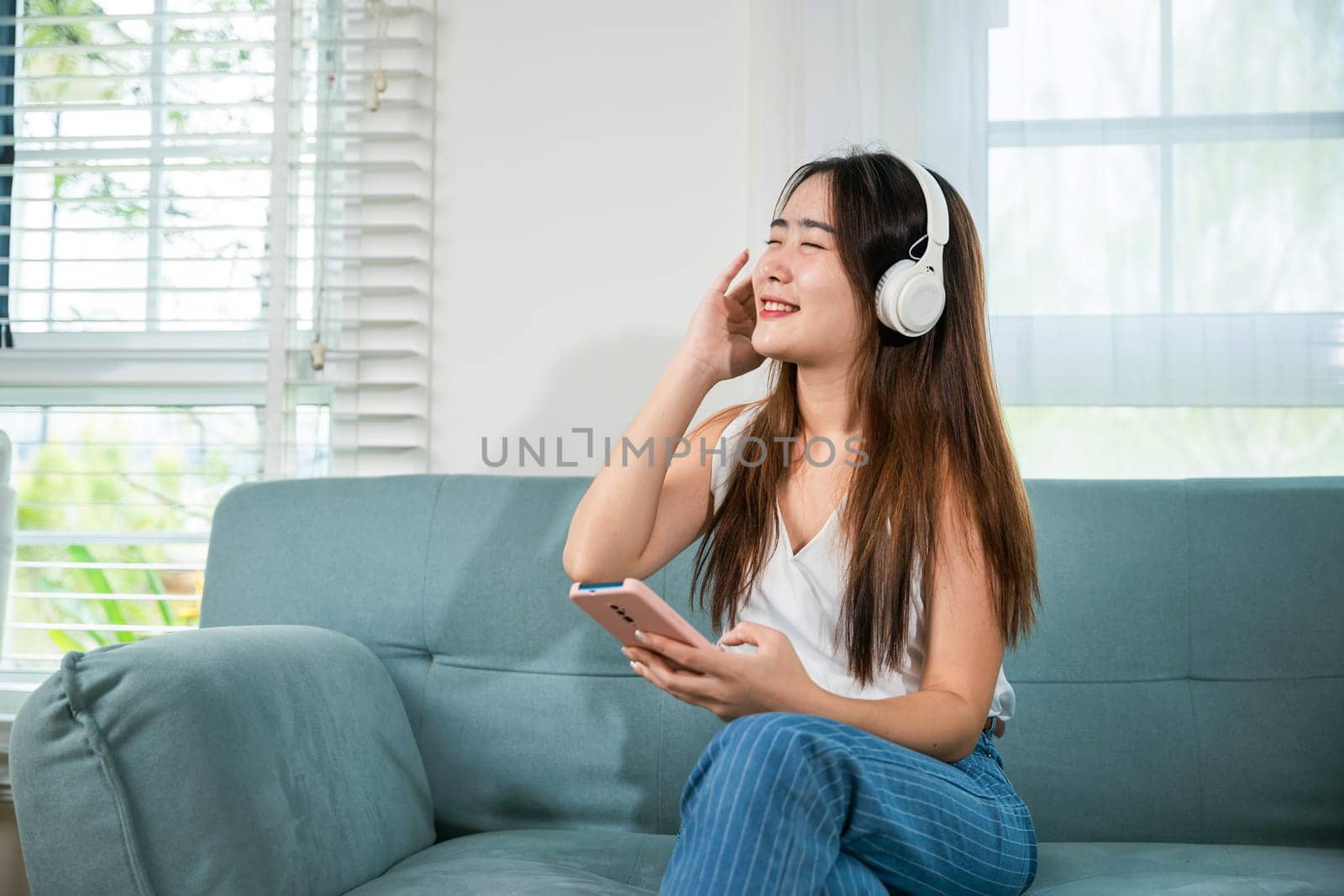 Asian young woman wearing headphones listening to music with smart mobile phone and relaxing while sitting on sofa in living room at home, female wear earphone listen music on smartphone