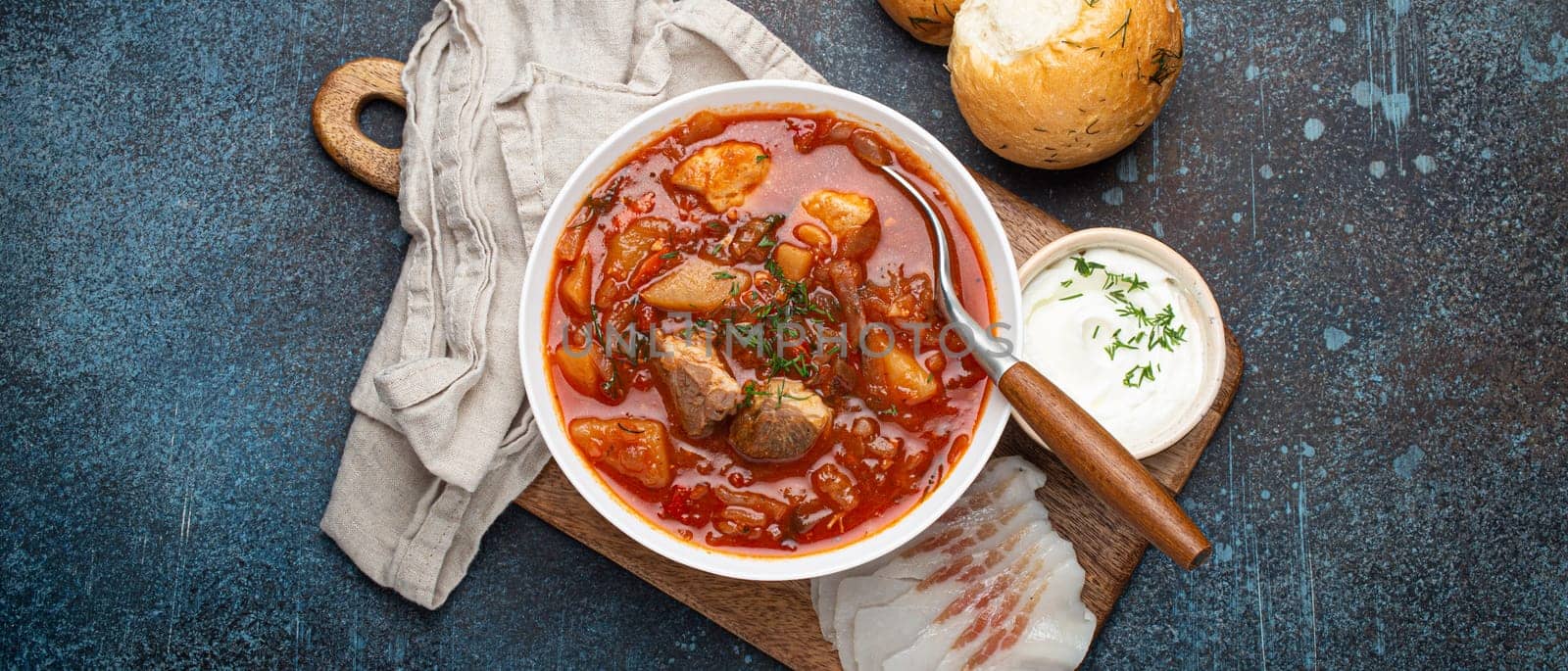 Ukrainian Borscht, red beetroot soup with meat, in white bowl with sour cream, garlic buns Pampushka and salo slices on rustic stone background. Traditional authentic dish of Ukraine