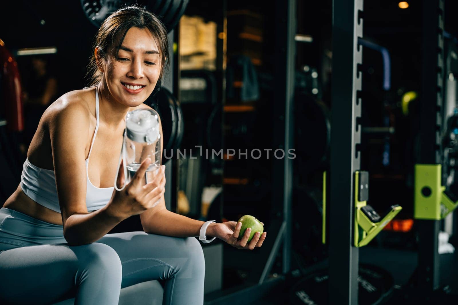 Asian girl holds green apple and water bottle in a gym, reminding viewers of the importance of balanced diet and hydration for achieving healthy lifestyle. Healthy fitness and eating lifestyle concept