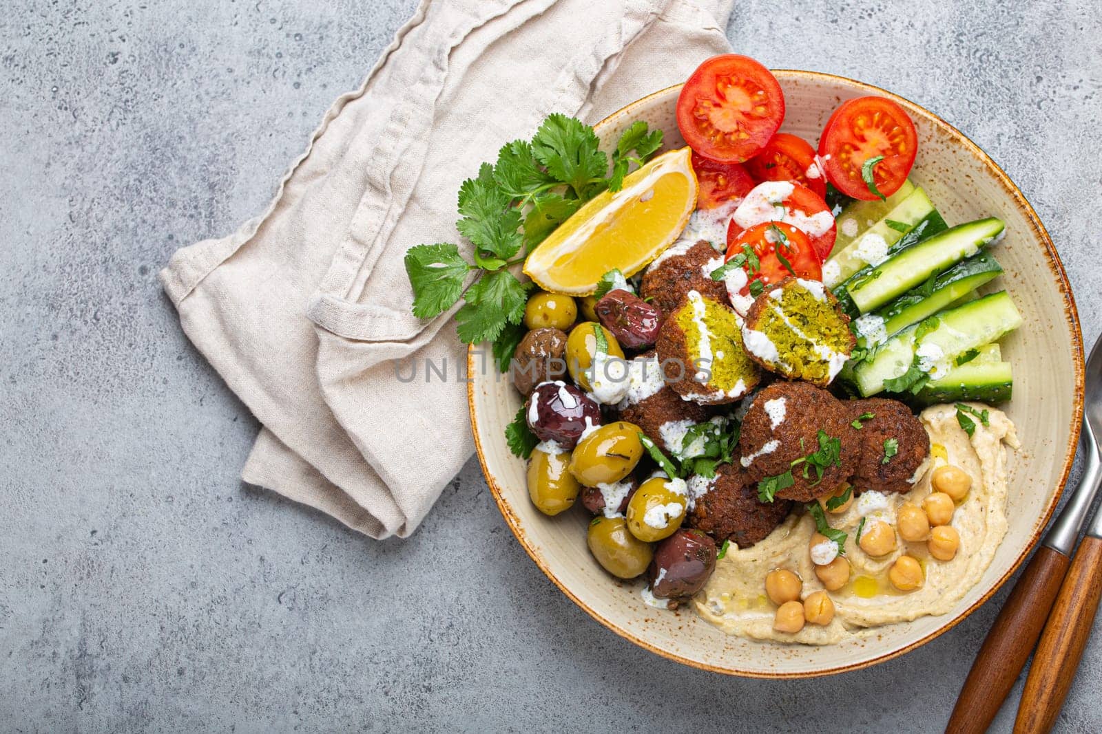 Falafel salad bowl with hummus, vegetables, olives, herbs and yogurt sauce. Vegan lunch plate top view on rustic stone background, healthy meal with falafel and veggies, copy space by its_al_dente