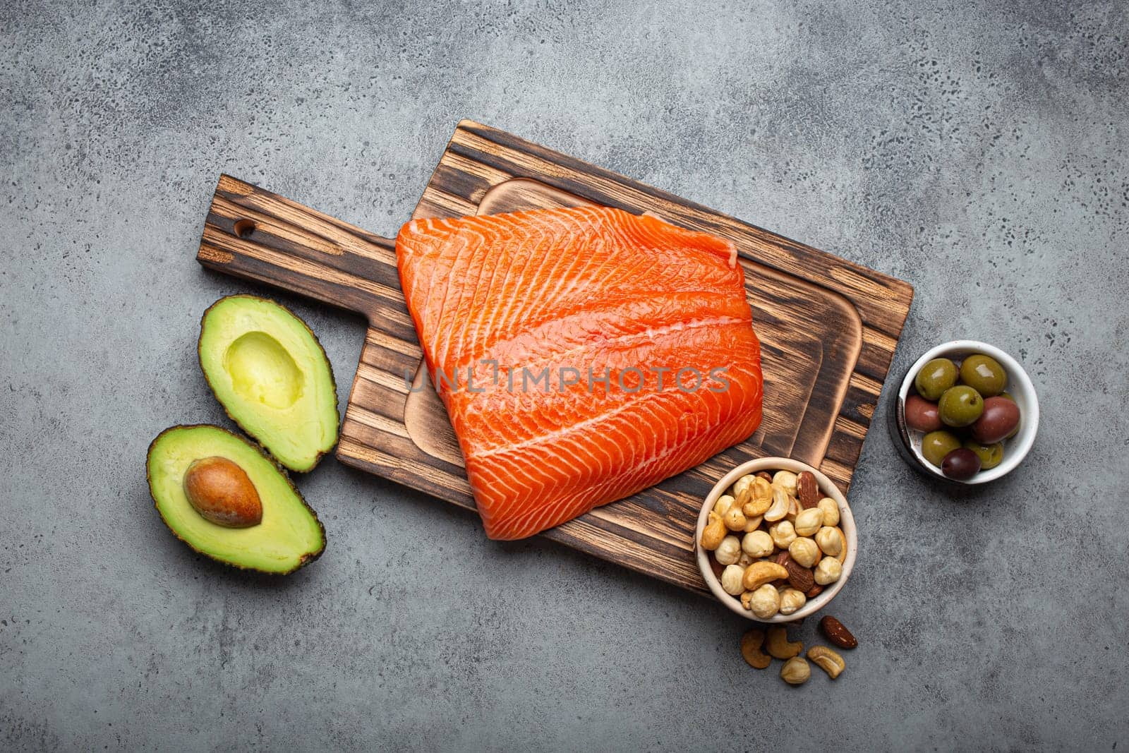 Food sources of healthy unsaturated fat: fresh raw salmon fillet, avocado, olives, nuts on cutting board, rustic stone background top view. Omega 3 and oil products, healthy nutrition and keto diet by its_al_dente