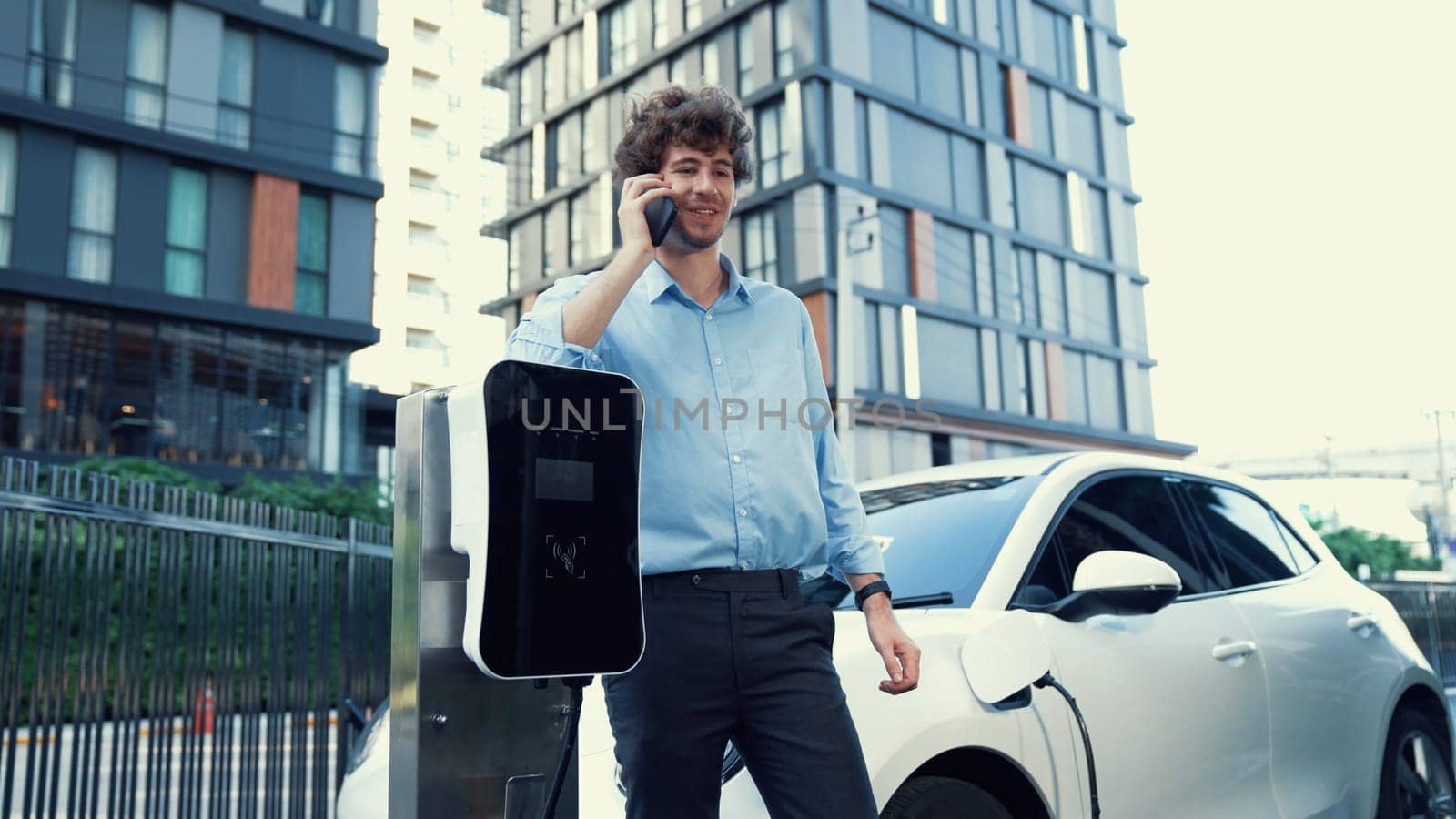 Suit-clad businessman with progressive ambition leaning on his electric vehicle while standing on a charging station with a power cable plug and a renewable energy-powered electric vehicle.