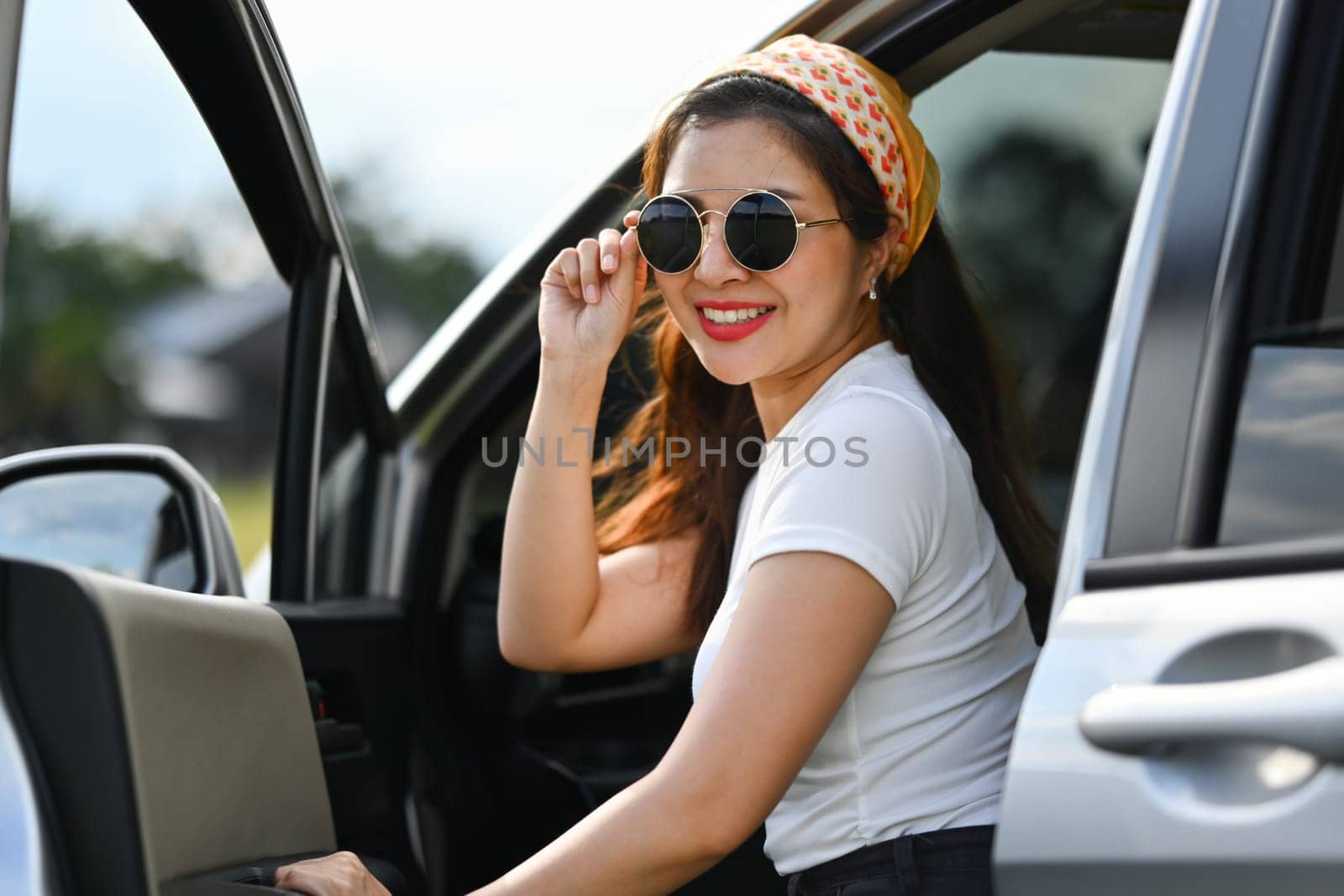 Confident female traveler wearing sunglasses sitting in car with open door. Travel and vacation concept.