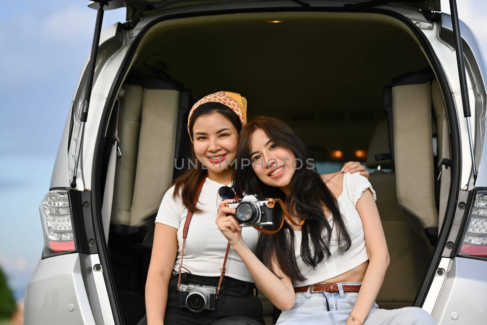 Two cheerful young women sitting on the open trunk of their car while taking a break from a road trip.